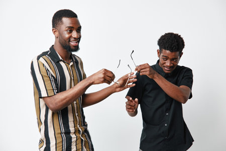 Two young black men with dark skin smiling and interacting while holding eyeglasses in casual...