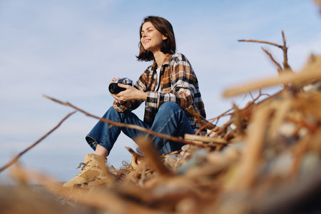 Woman sits outdoors among dried branches, wearing plaid shirt and jeans, holding a camera, enjoying...