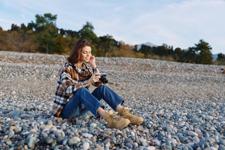 Outdoor woman photographer sitting on a rocky beach with a camera, wearing a plaid shirt and boots, enjoying a peaceful moment among pebbles and distant trees under a clear sky.の写真素材