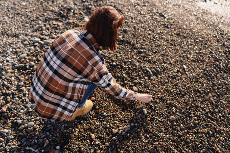 Person crouching on a pebble beach, wearing a plaid shirt, picking small stones as sunlight glints...