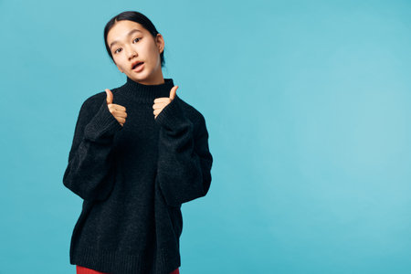 Young woman in black sweater with thumbs up gesture expressing approval and confidence against blue background. Casual style with relaxed pose and direct eye contact.の写真素材
