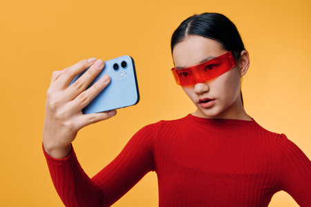 Young woman wearing red glasses taking selfie with smartphone on orange background. Female portrait with casual style for social media and digital photography content creation.の写真素材