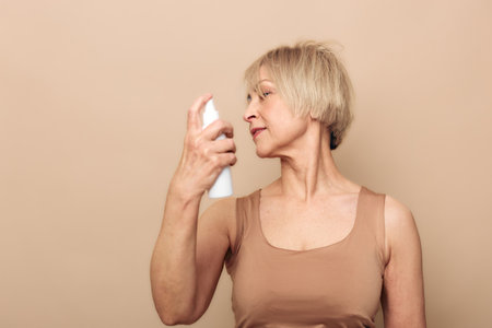 Mature woman with short blonde hair sprays mist on her face wearing beige tank top. Neutral background with soft lighting highlights skincare routine and self-care for aging skin.の写真素材