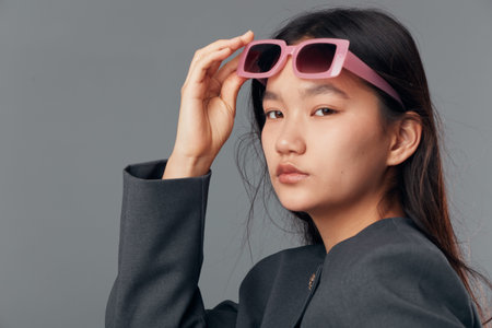 Young woman with long dark hair and serious expression wearing pink sunglasses and gray blazer posing on gray background in studio for fashion photography portrait.の写真素材