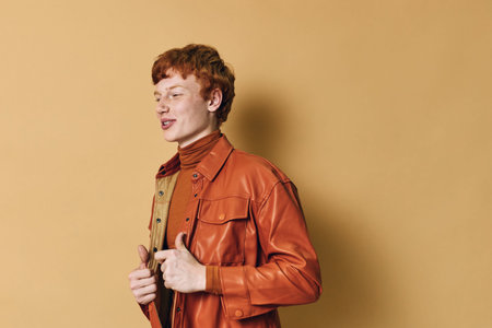 Young redhead man wearing orange leather jacket and turtleneck standing on beige background, posing with confident expression in casual style fashion portrait.の写真素材