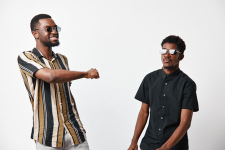 Two African American men with dark skin wearing sunglasses greeting each other with elbow bump on white background. Casual style, friendship and social interaction concept.の写真素材