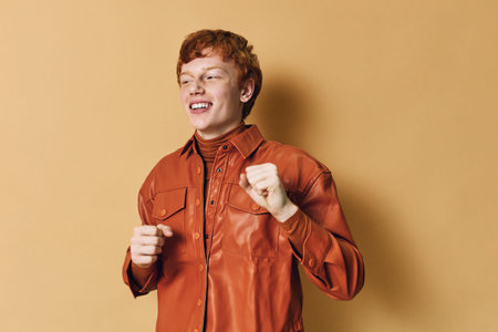 Young man with red hair wearing brown leather jacket smiling and dancing against plain background. Happy expression, casual style, youth culture and positive energy.の写真素材