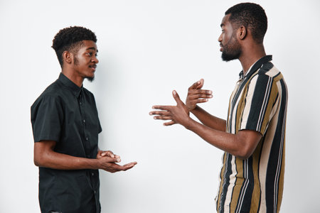 Two black men with dark skin having a conversation standing against white background. Casual clothing, communication, discussion, friendship, interaction, young adults, studio portrait.の写真素材