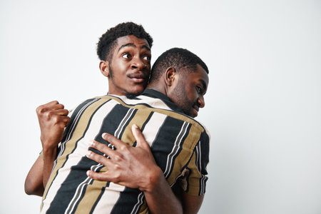 Two black men hugging each other with joy and excitement on white background. African ethnicity, dark skin, friendship and celebration with casual striped shirt.の写真素材