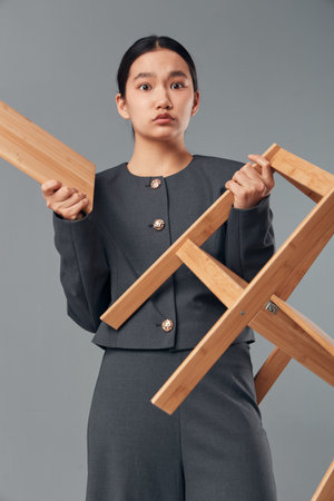 Woman with focused expression holding wooden chair frame in hands, dressed in dark formal outfit isolated on grey background for craftsmanship concept and furniture making.の写真素材