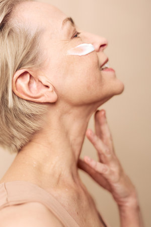 Mature woman with short blonde hair applies face cream on cheek, smiling gently. Close-up on beige background. Concept of skincare, aging, beauty, and self-care routine.の写真素材
