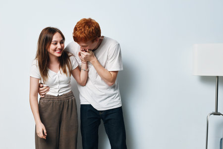 Young couple laughing and showing affection in casual clothes standing near white wall with lamp inside room, happy together and enjoying moment.の写真素材