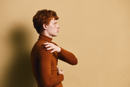 Profile of young man with red hair wearing brown turtleneck sweater standing against beige wall with serious expression and crossed arms.の写真素材