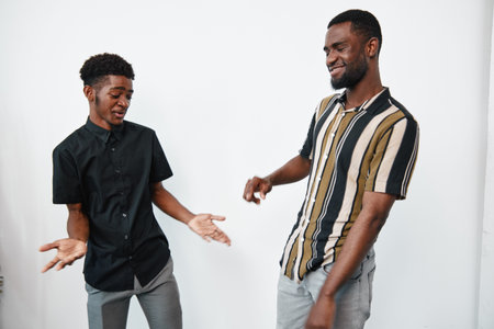 Two young black men with dark skin smiling and dancing together in casual clothes against white background in studio. Friendship and joy with happy emotions and movement.の写真素材