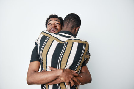 Two young black men with dark skin hugging each other in striped shirts, showing friendship and support in a close embrace against a white background.の写真素材