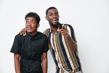 Two African American men with dark skin standing together on white background one pointing and smiling while the other looks surprised and thoughtful.の写真素材