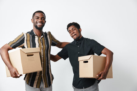 Two young black men holding cardboard boxes for moving or delivery, smiling and standing close together on white background, casual clothing and friendship.の写真素材