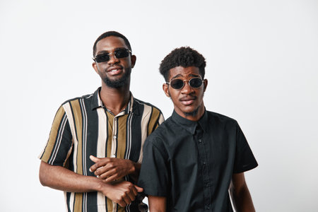 Two young black men with dark skin wearing sunglasses and casual shirts posing together on white background. Friends, style, fashion, confidence, portrait, studio, diversity.の写真素材