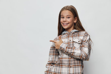 Teenage girl with long brown hair smiling and pointing to side wearing a checkered shirt on light background. Happy young female student with casual style and confident pose.の写真素材