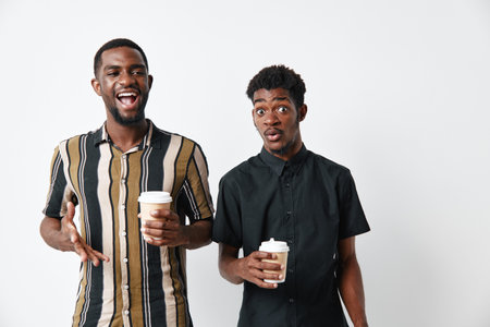 Two young black men with dark skin holding coffee cups and talking together in casual clothes on white background. Friends, communication and lifestyle with happy expressions.の写真素材