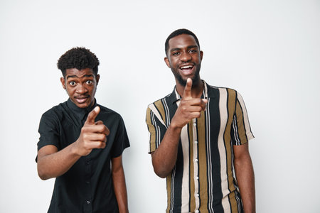 Two african men with dark skin pointing at camera smiling and standing together in casual clothes on white background. Young black males with happy expressions and friendly gesture.の写真素材