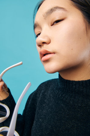 Woman with clear skin and natural look wearing black sweater holding white sunglasses close to face on blue background for fashion and lifestyle portrait.の写真素材