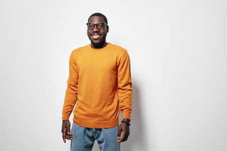 African American man with dark skin wearing orange sweater and glasses smiling standing against white background in casual outfit and relaxed pose.の写真素材
