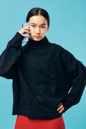 Young woman wearing black sweater with hand on hip talking on smartphone with serious expression against blue background. Casual style, technology and communication concept.の写真素材