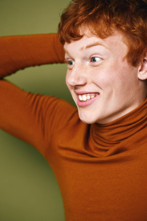Young man with red hair wearing brown turtleneck smiling looking sideways with hand behind head against green background. Emotion, casual, portrait, happiness, cheerful young adult.の写真素材