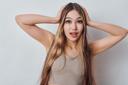 Young woman with long hair and surprised expression holding head with hands on white background, casual sleeveless top and natural makeup closeup portrait.の写真素材