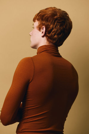 Profile view of a person with short red hair wearing a brown turtleneck top against a beige background. The subject is facing left with folded arms, showing a calm stance.の写真素材