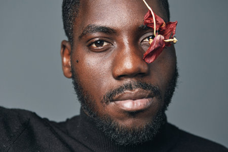 African man portrait with dried flower on face in creative conceptual visual style. Closeup of black male model with beard and serious expression on gray background for artistic photography.の写真素材