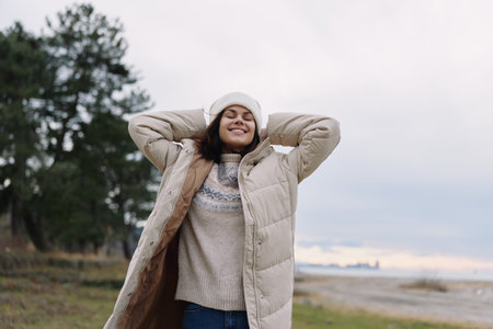 Woman outdoors wearing a warm coat and knit sweater, smiling with hands behind head, winter landscape and soft light, cozy fashion moment in chilly air and scenery.の写真素材