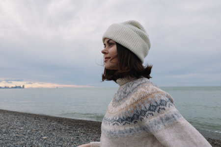 Woman in a knitted sweater and beanie on a pebbled beach with the ocean backdrop and a cloudy sky, capturing a calm outdoor momentの写真素材