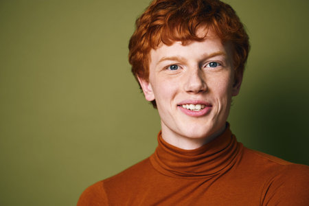 Young man with red hair and freckles smiling wearing orange turtleneck sweater on green background. Closeup portrait with natural expression and casual style in studio lighting.の写真素材