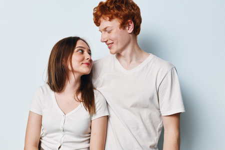Young couple standing against light background looking at each other smiling casual clothing together. Man and woman with red hair and brown hair posing and showing affection.の写真素材