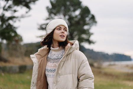 Woman in a warm winter jacket and knit sweater standing outdoors in an open field, wearing a beanie, gazing upward with soft natural light and distant trees.の写真素材