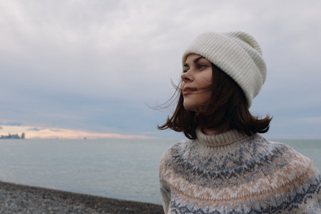 Portrait of a woman in a knit sweater and beanie by the sea, windblown hair, calm coastline and muted skies, showcasing outdoor fashion and serene coastal mood.の写真素材