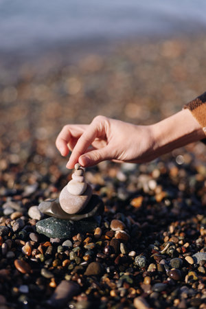 Stone balancing on a beach with a hand placing smooth rocks into a small stack among pebbles and sand, calm focus and meditation in a natural outdoor settingの写真素材