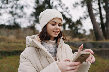 Woman in a warm coat and beanie outdoors, using a smartphone, relaxed and focused, capturing a candid moment in a chilly park setting during late autumnの写真素材