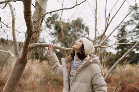 Woman in a beige winter coat and beanie reaches a branch in a bare tree landscape, capturing calm outdoor moments and a quiet seasonal mood during a cool day.の写真素材