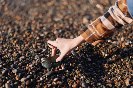 Closeup of a hand picking a dark stone from a bed of pebbles on a beach, showcasing natural texture and tactile detail in an outdoor setting.の写真素材