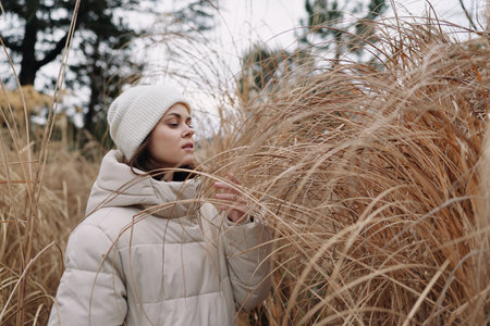 Winter, woman in a puffy coat and knit hat stands amid tall dried grasses in a quiet field, capturing serene outdoor moments, soft textures, and calm natural scenery.の写真素材