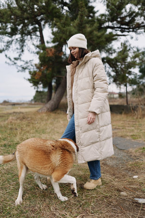 Woman in a warm puffer coat and beanie outdoors with a dog on a leash in a park, calm natural setting with trees and grass, candid moment of companionship and explorationの写真素材