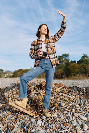 Adventure outdoor woman posing with camera on rocky terrain, casual plaid shirt and jeans, celebrating exploration under a clear sky with a raised handの写真素材