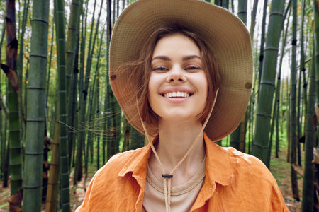 Woman smiling portrait in straw hat among bamboo forest, outdoor nature scene with casual orange shirt, cheerful traveler enjoying tropical greenery and peaceful relaxed mood.の写真素材
