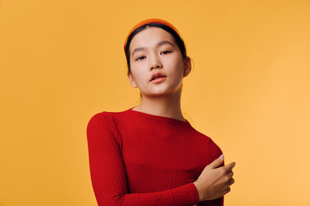Young woman wearing red sweater and headband posing with hand on shoulder against orange background for casual fashion portrait and style concept.の写真素材