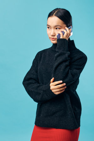 Young woman in black sweater talking on smartphone against blue background. Casual wear, technology use and communication with thoughtful expression and standing posture.の写真素材