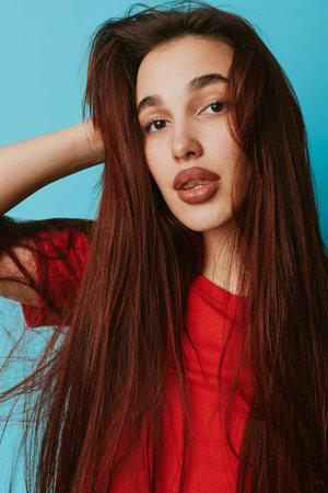 Young woman with long brown hair wearing red shirt posing with hand behind head against blue background. Portrait of female model with natural makeup and confident expression.の写真素材