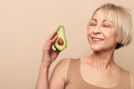 Smiling mature woman in beige tank top holds fresh avocado half near face with closed eyes. Concept of healthy lifestyle, natural nutrition, wellness, and organic food benefits for skin.の写真素材